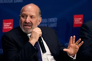A BALDING MAN WITH A BEARD AND MOUSTACHE IN A SUIT GESTURES WHILE HOLDING A MICROPHONE