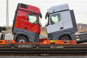 A red semi-truck and a white semi-truck stacked on a train chassis facing each other head-on,