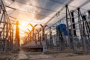 Rows of Y-shaped metal power transformers with wires criss-crossing the tops of each unit, set against a cloudy sunset