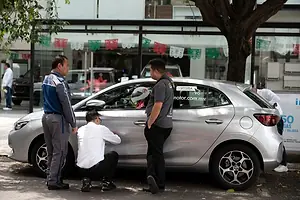 SEVERAL MEN CLUSTER AROUND A SILVER CAR, ONE IN A WHITE SHIRT CROUCHING DOWN