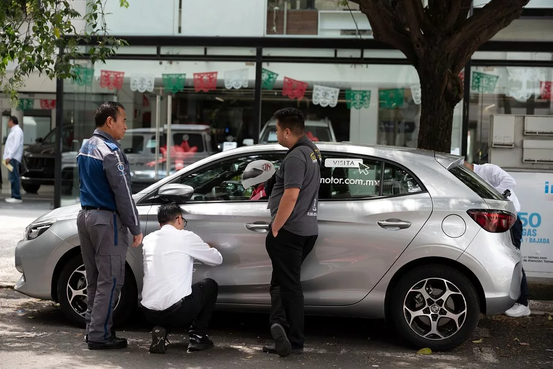 SEVERAL MEN CLUSTER AROUND A SILVER CAR, ONE IN A WHITE SHIRT CROUCHING DOWN