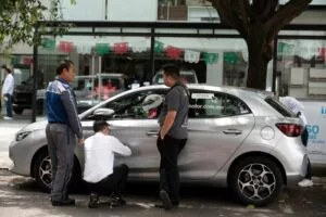 SEVERAL MEN CLUSTER AROUND A SILVER CAR, ONE IN A WHITE SHIRT CROUCHING DOWN