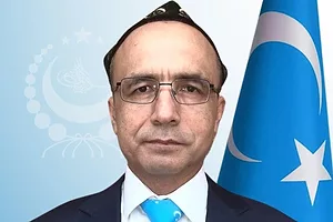 A man wearing glasses and a suit and light blue tie, standing in front of a blue flag with a crescent moon and a star