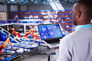 A man in a white dress shirt holding a laptop that has charts and graphs on a blue screen, in front of a warehouse of blue conveyor belts