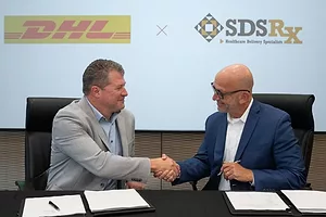 TWO SEATED MEN IN SUITS SHAKE HANDS IN FRONT OF COMPANY LOGOS