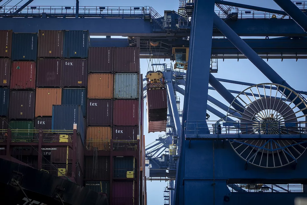 A tall stack of multi-colored shipping containers being removed one by one by a large blue shipping crane