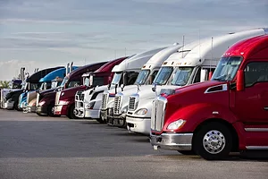 A row of semi-truck cabs, with a red truck in the foreground, parked next to four white trucks, two more red ones, and three blue ones