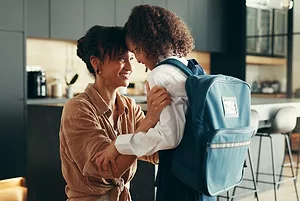 A WOMAN BENDS DOWN TO HUG A CHILD WEARING A BACKPACK