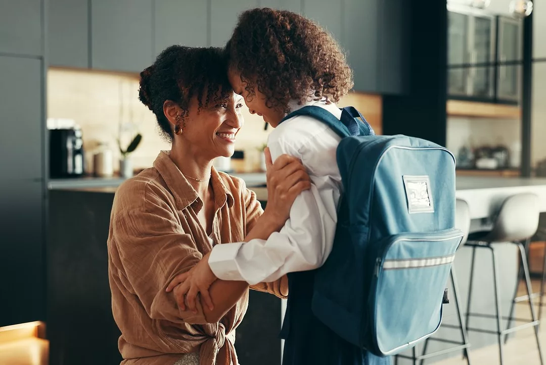 A WOMAN BENDS DOWN TO HUG A CHILD WEARING A BACKPACK