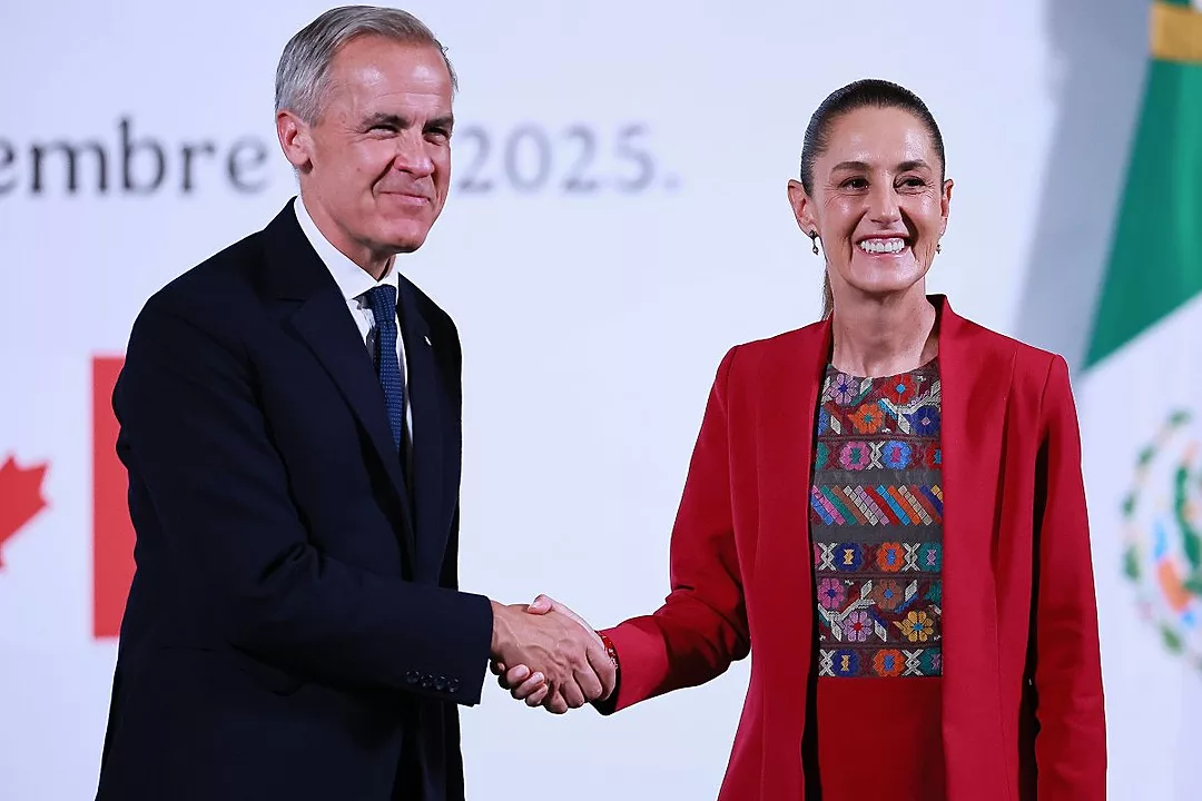 A MAN IN A DARK SUIT AND TIE SHAKES HANDS WITH A WOMAN IN A RED SUIT, BOTH TURNING TO SMILE FOR THE CAMERAS
