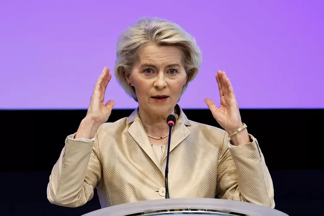A WOMAN IN A BEIGE SUIT AT A PODIUM RAISES BOTH HANDS TO MAKE A POINT