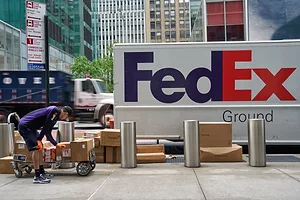 A WORKER WEARING THE FEDEX COLORS BENDS DOWN TO INSPECT A DOLLY LOADED WITH PARCELS ON A SIDEWALK IN FRONT OF A FEDEX TRUCK