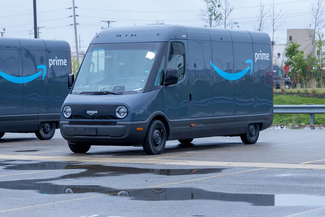 TWO BLUE DELIVERY VANS BEARING THE AMAZON LOGO SIT IN A WET PARKING LOT
