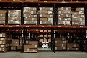 A WHITE ROBOT UNIT IS VISIBLE THROUGH A GAP IN WAREHOUSING STACKS