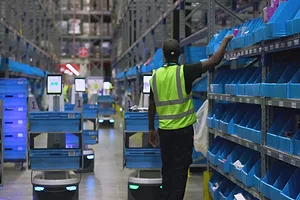 A WORKER IN A LONG WAREHOUSE AISLE REACHES UP TO A BLUE BIN, SURROUNDED BY WAIST-HIGH ROBOTIC UNITS