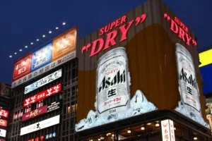 A large office building covered in display screens in neon lights, with a large wraparound screen on the corner advertising a silver can that reads "ASAHI"