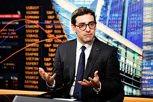 A MAN IN A DARK SUIT, TIE AND GLASSES, SITS RAISING HIS HANDS IN A QUESTIONING GESTURE