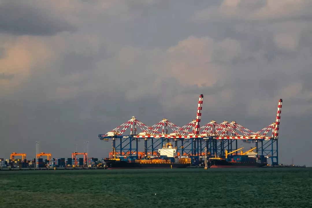 A row of red and white striped shipping cranes along a shoreline stacked with blue shipping containers