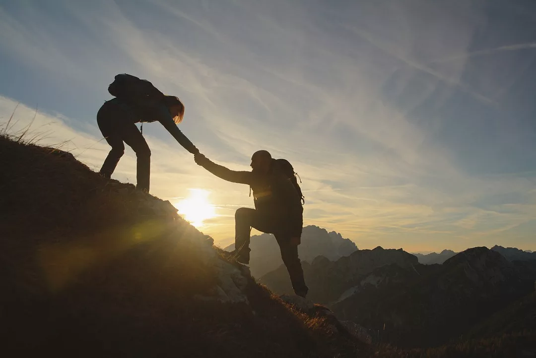 ON A STEEP HILL, ONE PERSON LINKS HANDS TO HELP ANOTHER UP
