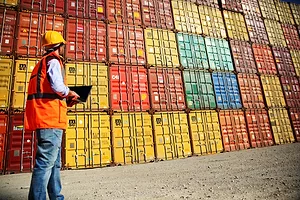 Multi-colored shipping containers stacked 10 rows across and 6 rows up, with a man in an orange safety vest standing in front