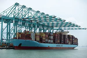 A blue container ship stacked with red and orange containers, docked underneath a row of blue shipping cranes on a cloudy day
