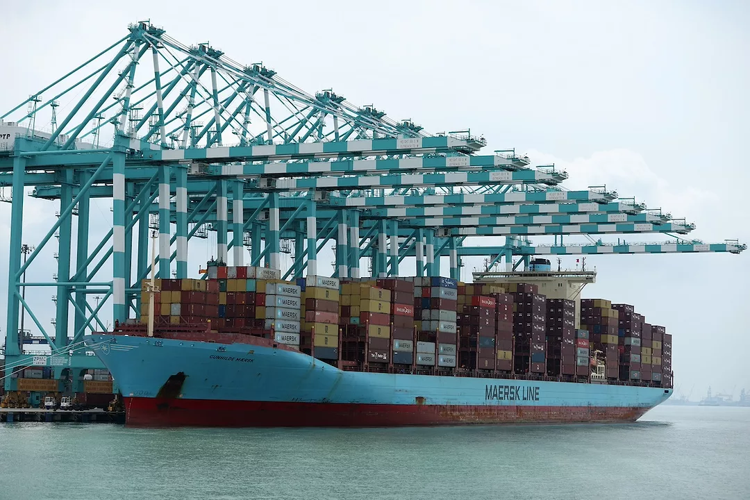 A blue container ship stacked with red and orange containers, docked underneath a row of blue shipping cranes on a cloudy day