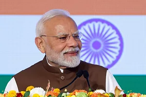 A WHITE HAIRED MAN WITH A WHITE BEARD WEARING A WHITE SHIRT AND A DARK NEHRU JACKET SMILES IN FRONT OF THE FLAG OF INDIA