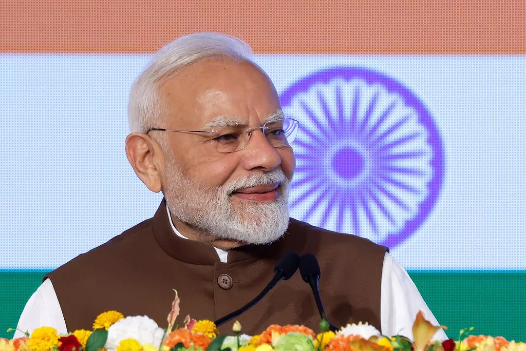 A WHITE HAIRED MAN WITH A WHITE BEARD WEARING A WHITE SHIRT AND A DARK NEHRU JACKET SMILES IN FRONT OF THE FLAG OF INDIA