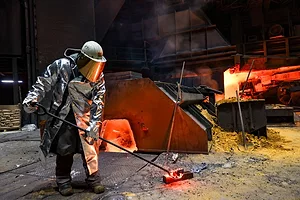 A WORKER IN HEAVY PROTECTIVE GEAR HANDLES A LONG POLE WITH A VESSEL OF MOLTEN METAL AT THE END, LIT BY THE GLOW OF A FURNACE