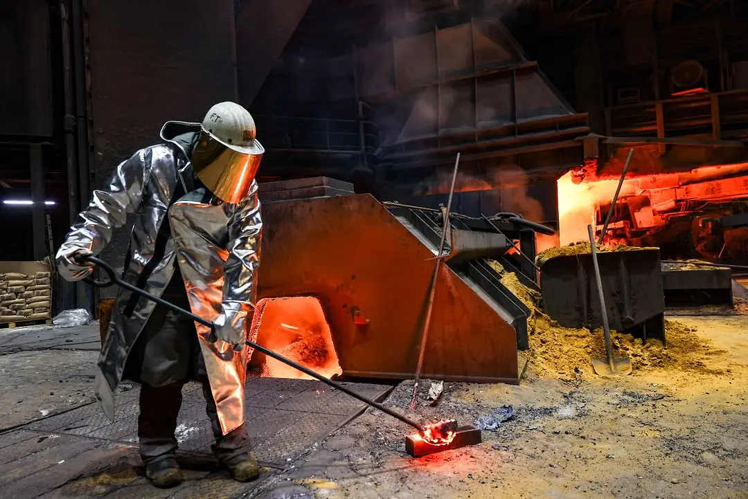 A WORKER IN HEAVY PROTECTIVE GEAR HANDLES A LONG POLE WITH A VESSEL OF MOLTEN METAL AT THE END, LIT BY THE GLOW OF A FURNACE