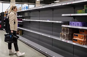 A SHOPPER IN A PONY TAIL AND SNEAKERS HOLDING A SHOPPING BASKET OBSERVES NEAR-EMPTY SHELVES IN A SUPERMARKET