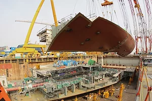 A rusted orange portion of a ship's hull dangled over a shipyard filled with metal platforms and surrounded by cranes