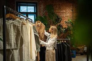 A SHOPPER WITH LONG BLOND HAIR PULLS A JACKET FROM A RACK OF CLOTHES IN A HIGH-END, TRENDY STORE