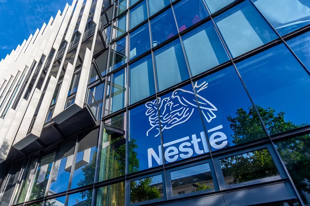 An office building with clear glass windows and "NESTLE" printed in white lettering on the lower right, below an image of three birds in a nest