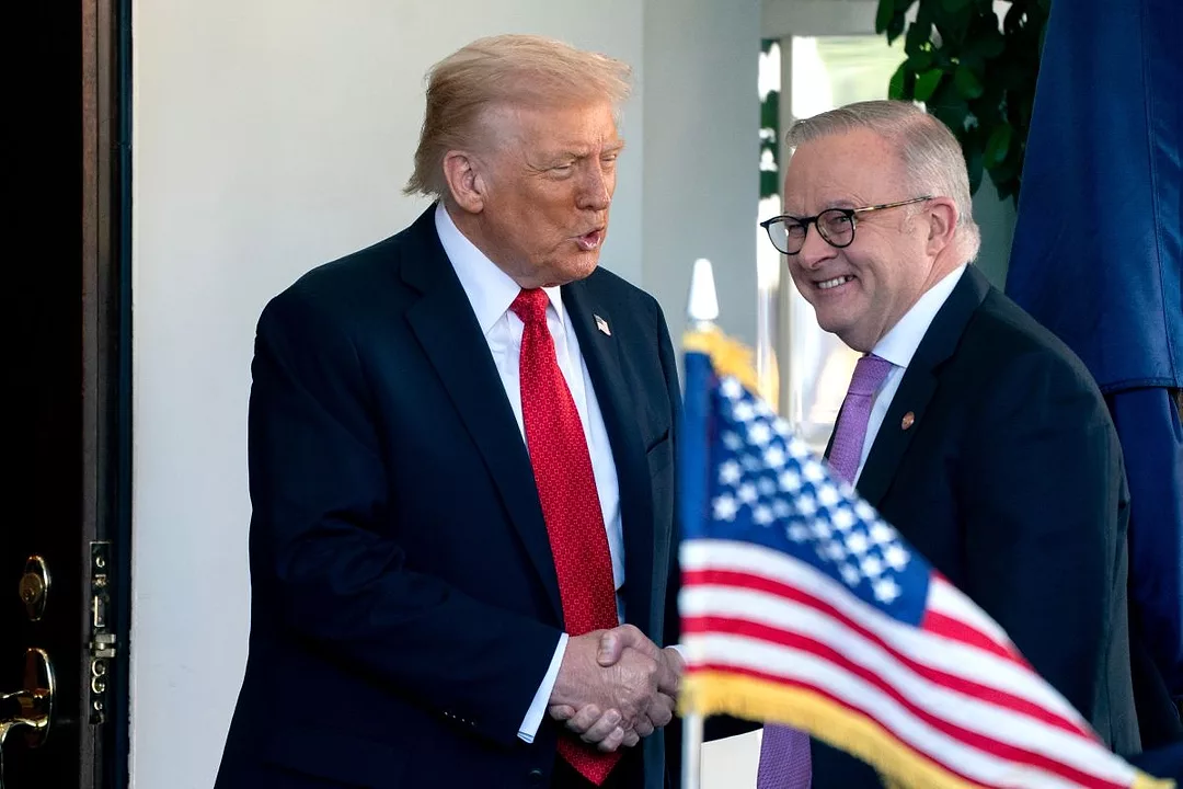 A TALL MAN IN A BLUE SUIT AND RED TIE SHAKES HANDS WITH A GRAY-HAIRED MAN IN A SUIT AND GLASSES