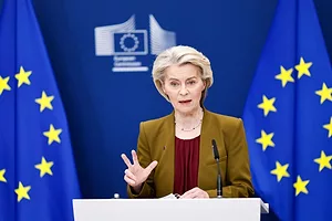 AN ELEGANT OLDER WOMAN IN A BROWN JACKET SPEAKS FROM A PODIUM IN FRONT OF BLUE AND GOLD EU FLAGS