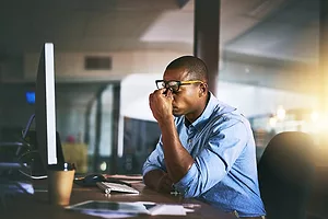 A MAN AT A COMPUTER PINCHES THE TOP OF HIS NOSE UNDER HIS SPECTACLES, IN FRUSTRATION