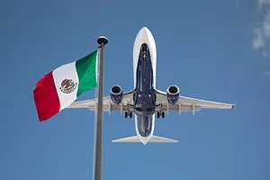 A PLANE FLIES THROUGH BLUE SKIES PAST A FLAG OF MEXICO