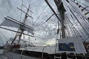 A large sailing vessel with two masts and a white sail, docked at a port on a cloudy day