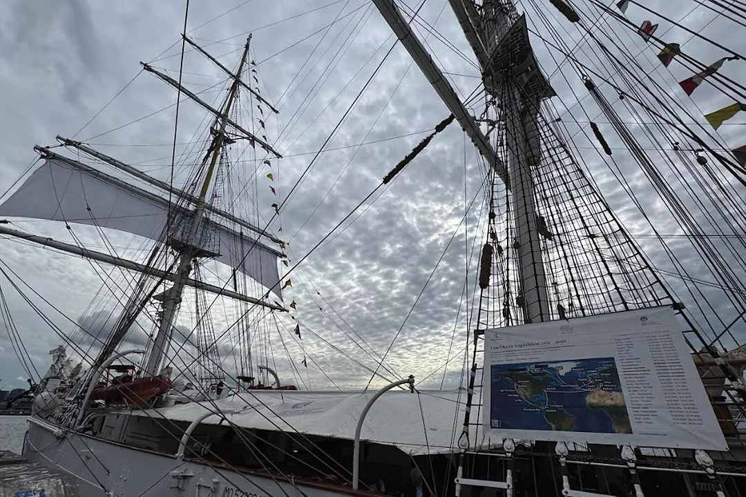 A large sailing vessel with two masts and a white sail, docked at a port on a cloudy day