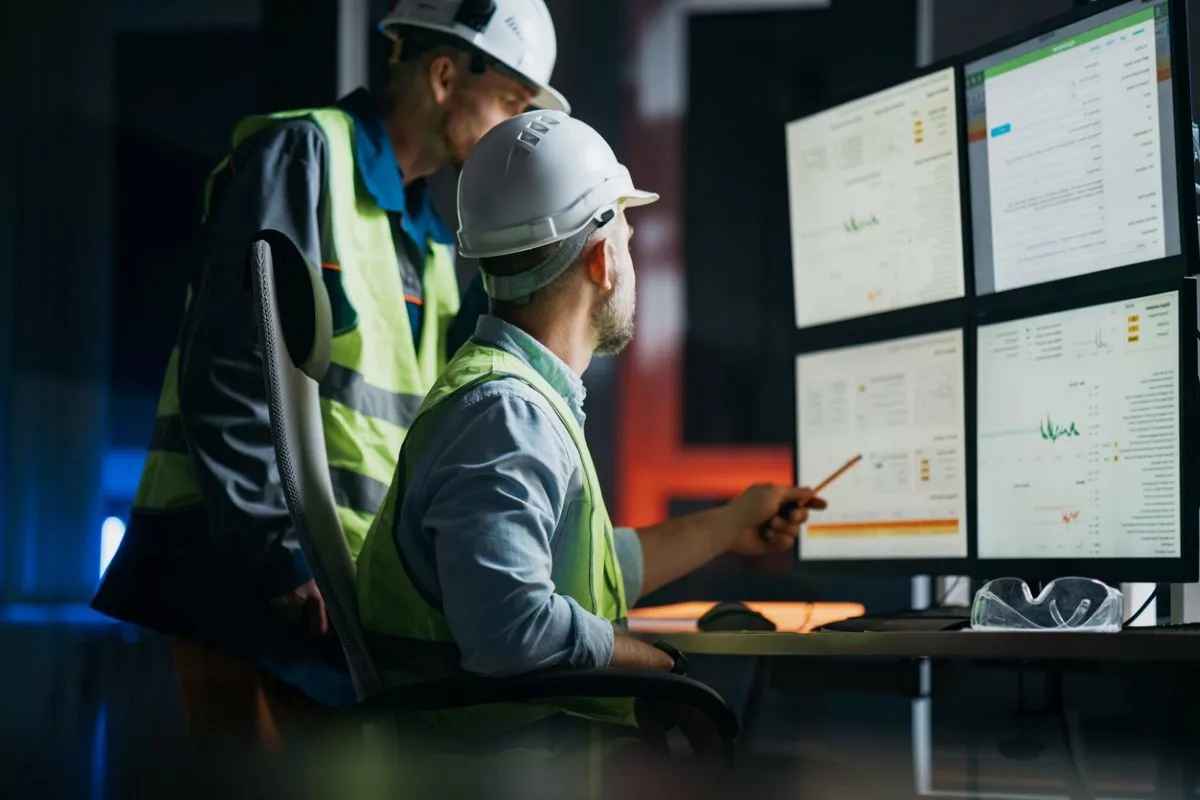 TWO WORKERS IN HI-VIS VESTS AND HARDHATS CONSULT A BANK OF COMPUTER SCREENS
