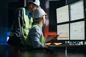 TWO WORKERS IN HI-VIS VESTS AND HARDHATS CONSULT A BANK OF COMPUTER SCREENS