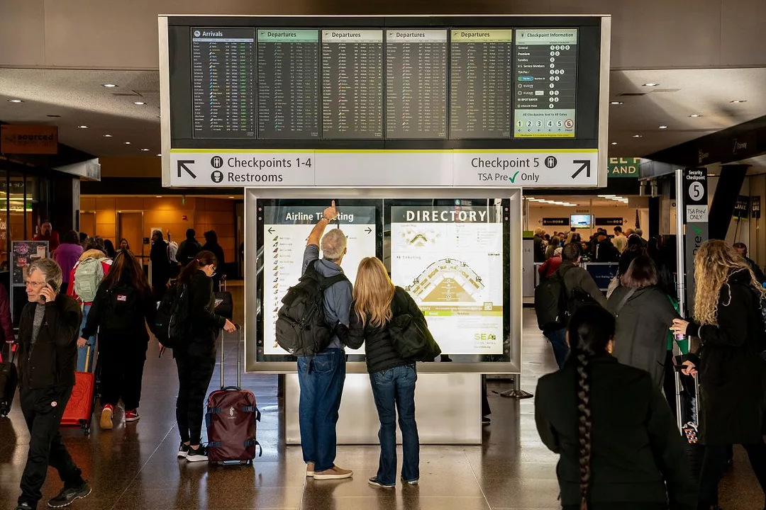Travelers look at an arrivals and departures board at a busy airport.