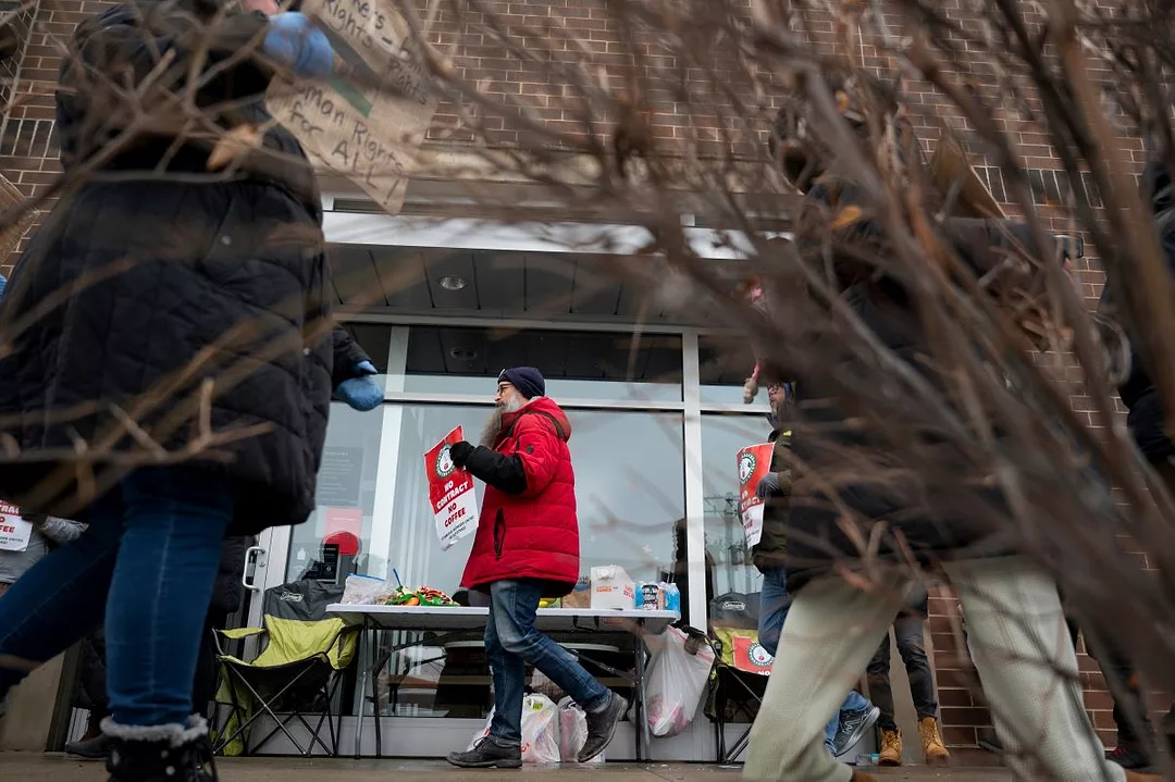 A PERSON IN A BRIGHT RED DOWN COAT HOLDS A PLACARD OUTSIDE A STOREFRONT