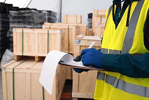 A man in a yellow safety vest holding a clipboard, and standing in front of stacks of wooden crates