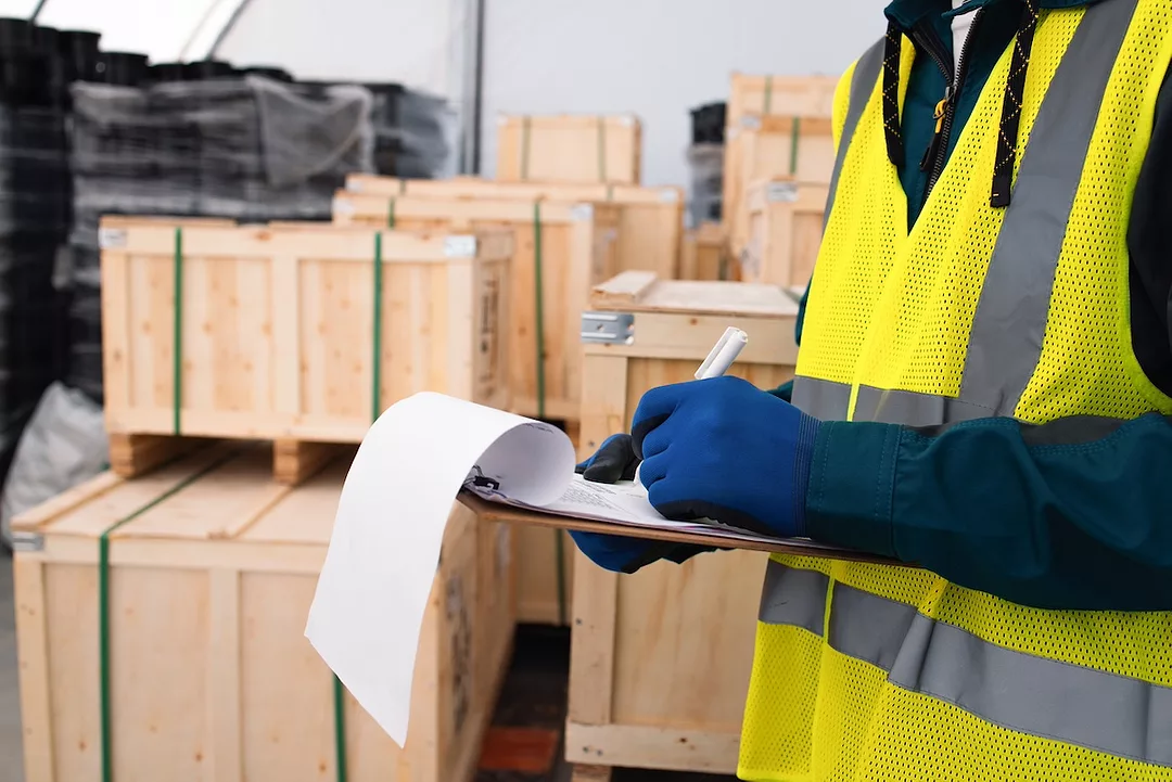 A man in a yellow safety vest holding a clipboard, and standing in front of stacks of wooden crates