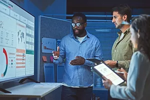 THREE COLLEAGUES, ONE SEATED, TWO STANDING, CONSULT EARNESTLY AROUND A BANK OF COMPUTER SCREENS