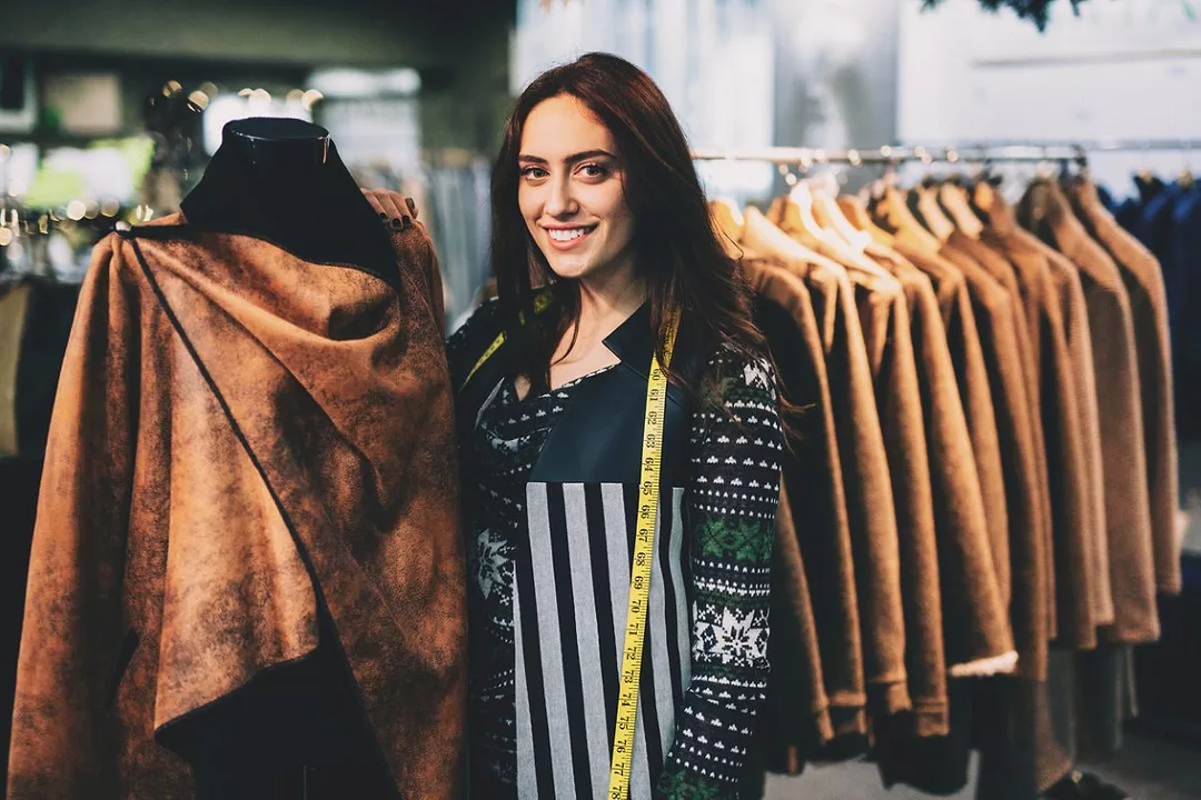 A WOMAN WITH A MEASURING TAPE ROUND HER SHOULDERS STANDS SMILING IN FRONT OF A RACK OF CLOTHING