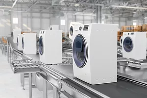 A line of white front-load washing machines on a conveyer belt in a large warehouse