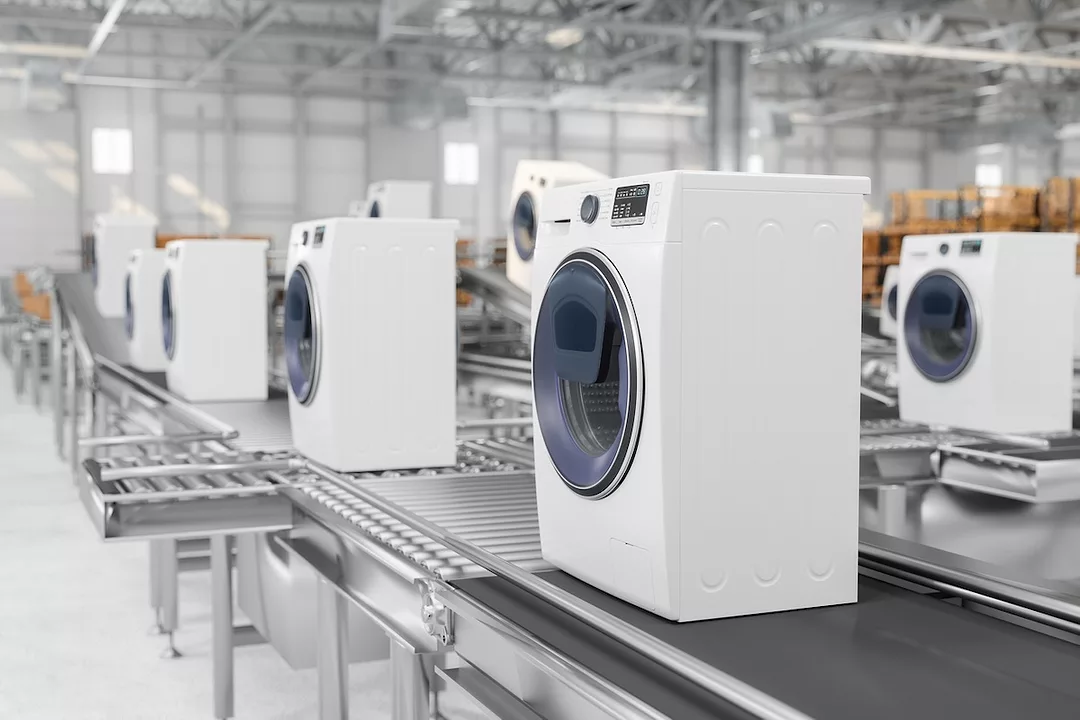 A line of white front-load washing machines on a conveyer belt in a large warehouse